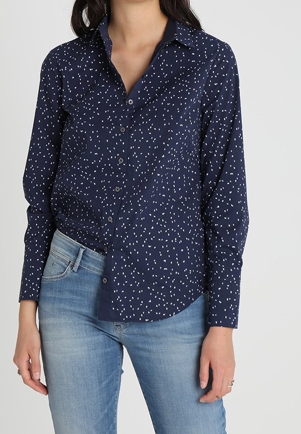 Chemise bleu marine à boutons avec un col à revers et un motif de petits points blancs éparpillés. Manches longues avec poignets repliés. Portée avec un jean bleu clair.