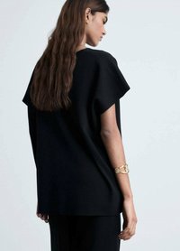 Černý oversized top s volným střihem a širokými rukávy, vyrobený z měkké látky. Na pravém zápěstí se nachází zlatý náramek.