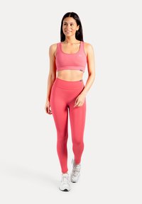 Roze sportbeha en leggings met hoge taille van een gladde, rekbare stof. Model draagt witte sportschoenen en staat op een neutrale achtergrond.