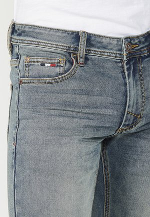 Jean en denim bleu clair avec une coupe slim, présentant des coutures jaunes, un petit patch multicolore sur la poche, et un design classique à cinq poches.