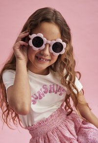 Rosa blommformade solglasögon med svarta linser. Vit t-shirt med "SUNSHINE" i rosa paljetter. Rosa mönstrad kjol med smockad midja.