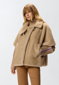 Cappa beige, morbida, in misto lana con un ampio colletto, bottoni a toggles e tasche laterali; presenta maniche a coste di un beige più chiaro.