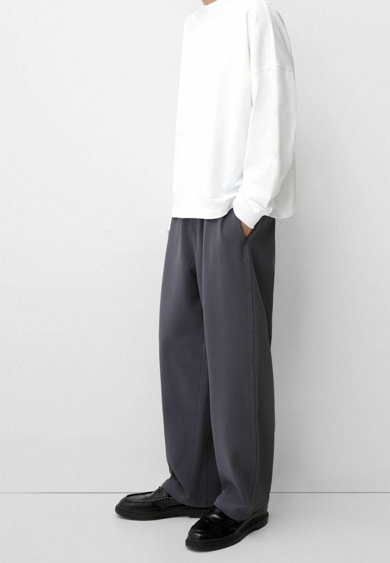 Weißes Langarmhemd mit lässiger Passform, kombiniert mit grauen weit geschnittenen Hosen. Das Outfit wird durch schwarze Schnürschuhe ergänzt.