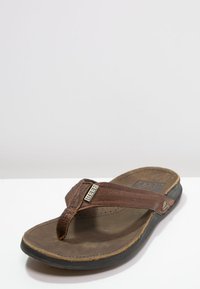 Brune lær flip-flop sandaler med perforerte stropper, en strukturert såle og gummisåle. Har en metallisk logo aksent på stroppen.