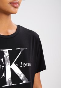 Svart bomull t-shirt med en stor vit grafik "CK" med marmorliknande textur och texten "in Jeans" nedanför. Korta ärmar och rundad halsdesign.