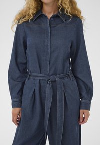 Ženska v temnodeminskem kombinezonu z dolgimi rokavi in ovratnikom, s sprednjim gumbom in zawičenim pasom iz blaga, ki stoji ob beli ozadju.