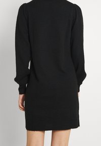 Une personne portant une robe en tricot noir à manches longues, vue de dos, se tenant contre un fond clair et uni.