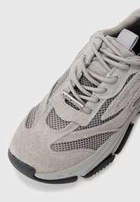 Gray athletic shoe featuring a mix of suede and mesh materials, laces, and a prominent black rubber sole. Notable silver hardware detail.