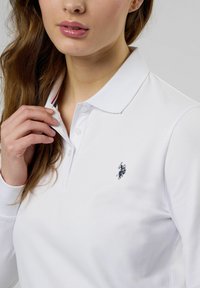 Sieviete pieskārās baltas, ar garām piedurknēm hvardes krekla apkaklei, kuram uz krūtīm ir mazs tumšs zirgs un jātnieks, fona ir vienkāršs.