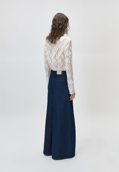 Γυναίκα με σγουρά μαλλιά που φοράει λευκό μπλουζάκι με μακρύ μανίκι από δαντέλα με λουλούδια και ψηλόμεσα τζιν με ευρύ πόδι σε σκούρο μπλε χρώμα, με την πλάτη στραμμένη.