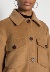 Brun ullkappa med krage, fyra stora fickor och mörkbruna knappar. Mjuk textur med skräddarsydd passform.