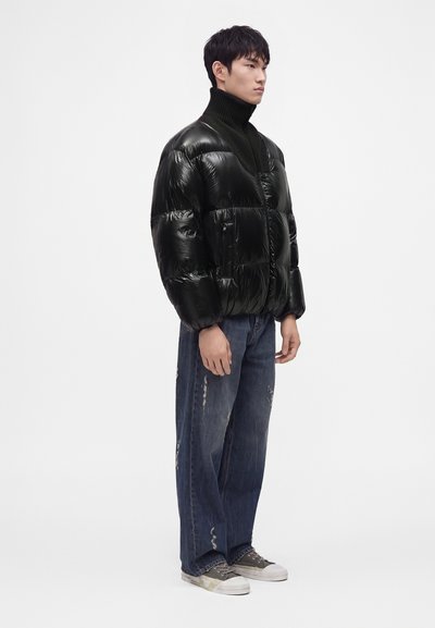 Jeune homme debout portant une veste matelassée noire brillante, un col roulé noir, un jean bleu large et des baskets basses grises sur fond blanc.