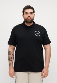 Juodas polo marškinėlis, pagamintas iš medvilnės, su apykakle, trumpomis rankovėmis ir apvaliu baltu logotipu kairiajame krūtinės kampe. Klasikinio fasono dizainas.