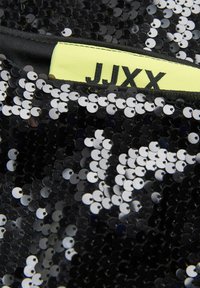 Crna tkanina s cirkonima koja sadrži okrugle cirkone, s vidljivom žutom etiketom urezanom s "JJXX" na crnoj traci.