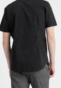 Man med kortärmad svart skjorta och grå strukturerade byxor, visad bakifrån mot en enkel bakgrund.