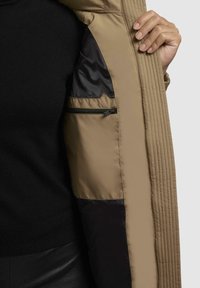 Beige gevoerde jas met een gequiltde textuur, zwarte voering en een ritszak. De stof is glad en lichtgewicht, met een relaxed fit.