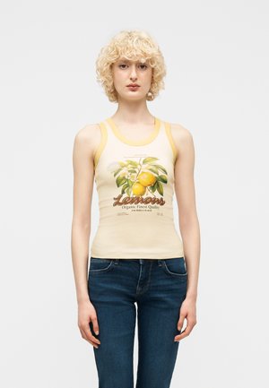 Personne blonde portant un haut sans manches à bord jaune avec un motif de citron et du texte, assorti à un jean bleu foncé, debout sur fond blanc.