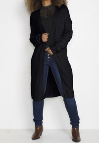 Długi czarny sweter z dzianiny z teksturowanym wzorem, otwarty przód i ściągacze, zestawiony z czarną bluzką i niebieskimi dżinsami. Brązowe botki na obcasie.