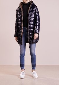 Marinblå glänsande pufferjacka med huva, med dragkedjestängning och ribbat innertyg. I kombination med tighta blå jeans och vita sneakers.