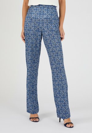 Femme portant un pantalon bleu taille haute, coupe droite, avec un motif floral géométrique et des talons ouverts bleu marine, debout sur fond blanc.