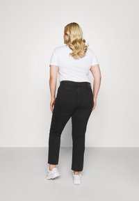Mujer con cabello largo y rubio, vestida con una camiseta blanca, jeans negros y zapatillas blancas, de pie de espaldas contra un fondo gris liso.