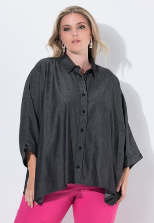 Donna bionda in una camicia grigia scuro oversize con bottoni e pantaloni rosa brillante, che indossa orecchini geometrici in argento, in piedi contro un muro chiaro.
