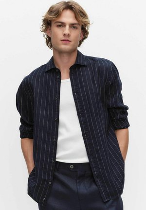 Młody mężczyzna o jasnobrązowych włosach, ubrany w granatową koszulę w prążki na białej koszulce, z rękami w kieszeniach, stoi na tle białego tła.