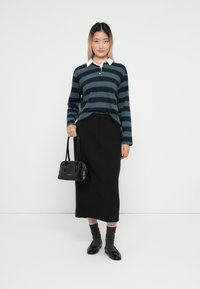Chemise rayée à manches longues en vert foncé et bleu marine, associée à une jupe midi noire, tenant un sac à main noir, portant des chaussettes grises et des ballerines noires.