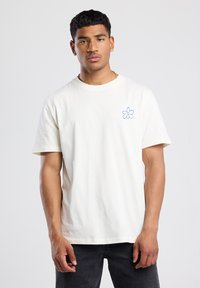 Katoenen t-shirt in off-white met korte mouwen en ronde halslijn. Heeft een blauw bloemenontwerp op de linkerborst.