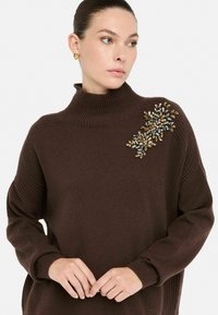 Brązowy sweter dzianinowy z wysokim kołnierzem i prążkowanymi mankietami, ozdobiony dekoracyjnymi zdobieniami w złotym, niebieskim i innych odcieniach na ramieniu.