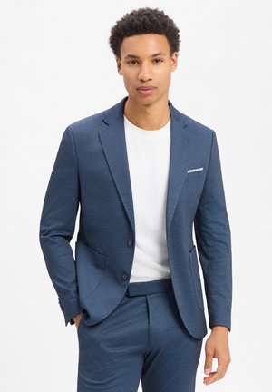 Junger Mann, der einen blauen Anzug mit einem weißen T-Shirt mit Rundhalsausschnitt trägt, Hände in den Taschen, steht vor einem schlichten weißen Hintergrund.