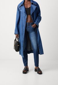 Denimlång kappa med breda slag, ihop med mörkblå jeans och svarta klacksandaler med blockklack; håller en liten svart handväska med guldaccenter.