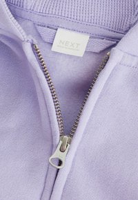 Fialová mikina na zip s metalovým zipem, ribovaným límcem a měkkým vnitřkem. Na štítku je nápis "NEXT, MADE IN PAKISTAN."