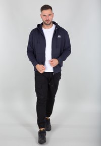Felpa con zip blu marina e tasche frontali, sopra una maglietta bianca. Pantaloni cargo neri con cerniere e orli risvoltati, abbinati a scarpe da ginnastica nere.