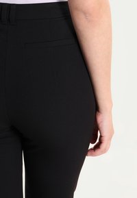 Närbild av en persons nedre rygg och höger arm som bär åtsittande svarta byxor med bakficka och en instoppad vit topp.