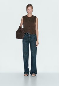 Haut sans manches en tricot marron avec un devant à boutons et deux poches, associé à un jean évasé bleu foncé. Le modèle tient un sac en suède marron.