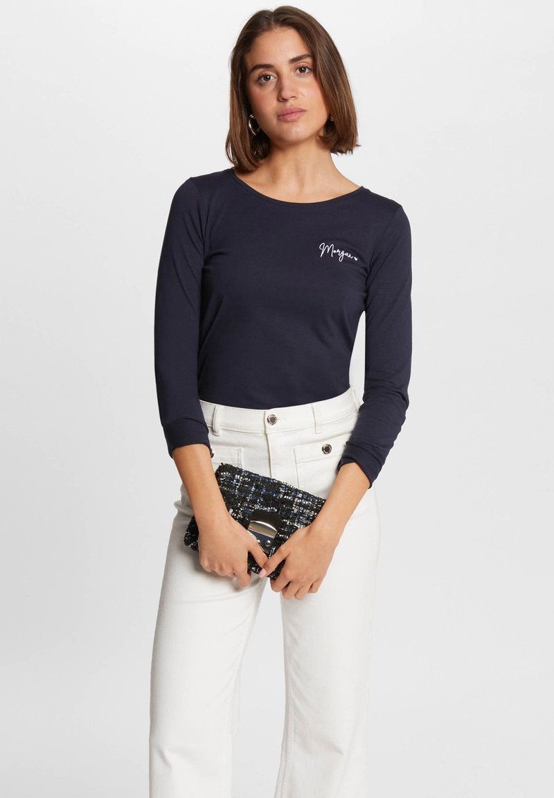 Dunkelblaustes Langarmoberteil mit rundem Ausschnitt und weißem Schriftzug-Logo; kombiniert mit hochgeschnittenen weißen Hosen und einer strukturierten schwarzen Clutch.