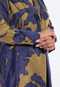 Blusa floral en oliva y marino. Cuenta con un escote en V, mangas largas con puño y botón, y un frente plisado. Tejido suave y texturizado.