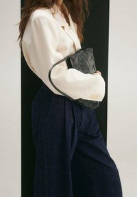 Una mujer con un blazer crema y pantalones de pierna ancha azul marino sostiene un bolso de cuero trenzado negro frente a un fondo en blanco y negro.