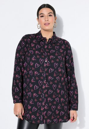 Kvinde med mørkt hår iført en sort langærmet skjorte med mønster af lyserøde hjerter og sløjfer, iført guld øreringe og ringe, stående foran en hvid baggrund.