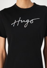 Black cotton t-shirt featuring a white holographic "Hugos" logo in cursive across the chest. Short sleeves and a classic crew neckline.