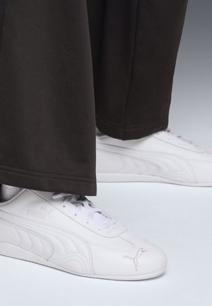 White Puma sneakers worn with black wide-leg pants, focusing on side and top view of shoes on a light gray surface.