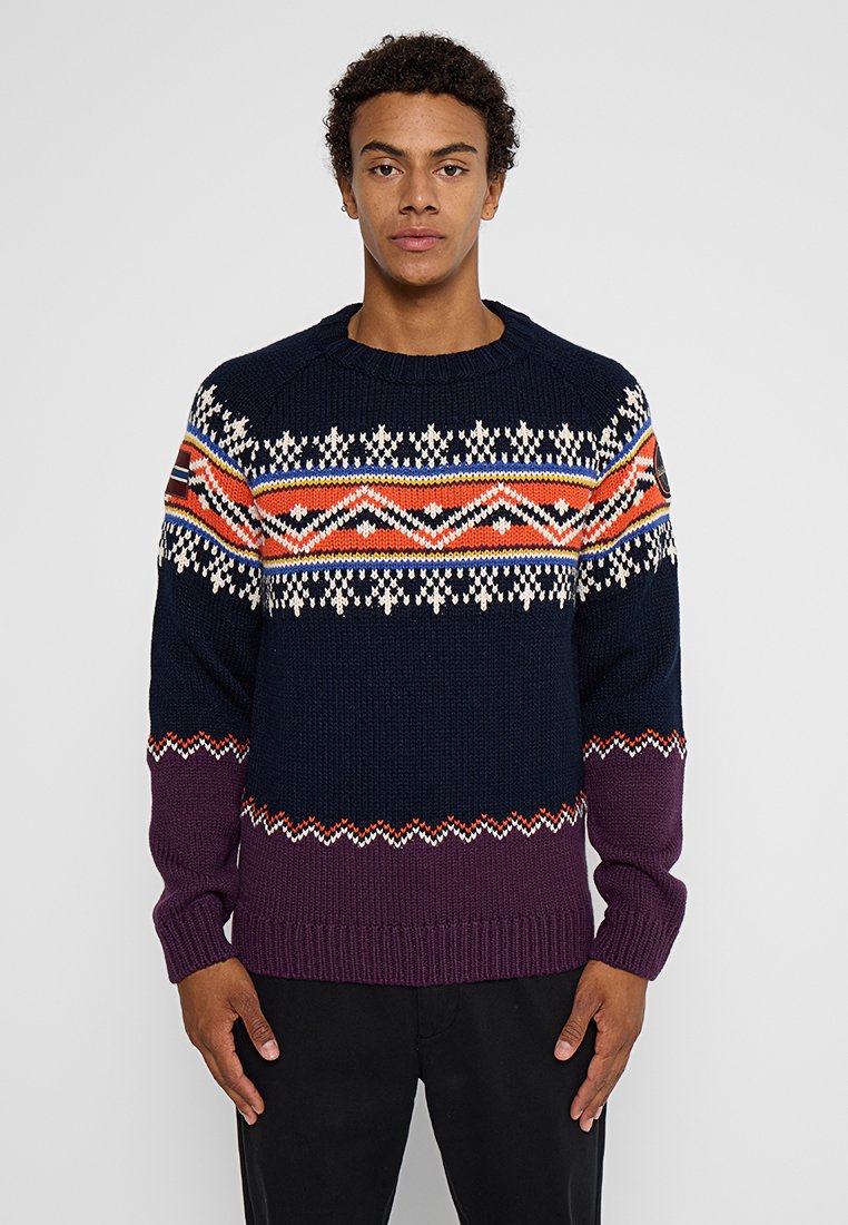 Jeune homme portant un pull en laine tricoté bleu marine et violet avec des motifs géométriques orange, blancs et bleus, se tenant devant un fond gris uni.