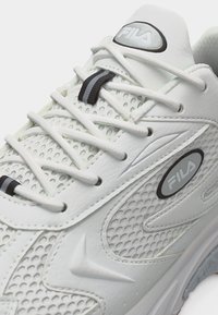Λευκό sneaker Fila με υφασμάτινα πάνελ με υφή, λευκά κορδόνια περασμένα από οπές και μαύρους-γκρι branded αναστολείς κορδονιών.