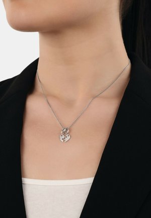 Collana d'argento con un pendente a forma di cuore caratterizzato da un design a puzzle, attaccata a una catena sottile, indossata sopra un blazer nero.