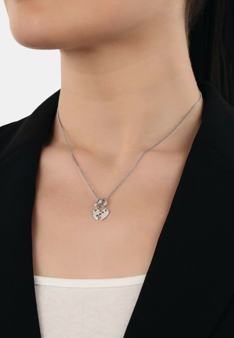 Collana d'argento con un pendente a forma di cuore caratterizzato da un design a puzzle, attaccata a una catena sottile, indossata sopra un blazer nero.
