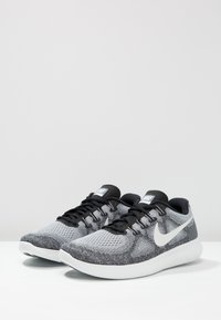 Un paio di scarpe da corsa Nike grigie e nere con suole bianche e loghi swoosh bianchi su una superficie bianca e sfondo grigio chiaro.