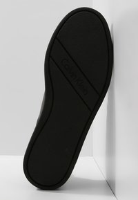 Semelle extérieure en caoutchouc noir avec surface texturée, présentant une bande horizontale avec le logo "Calvin Klein" dans un design épuré et minimaliste.