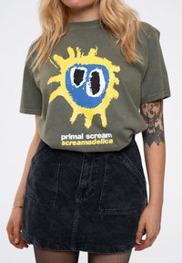 Olive green cotton t-shirt with a yellow and blue graphic design. Paired with a black denim skirt featuring front pockets and a textured finish.