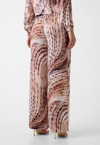 Faltenweite Hosen aus einem glatten, leichten Stoff mit einem beige-braunen wirbelnden Muster. Die Säumen sind sauber und glatt verarbeitet.